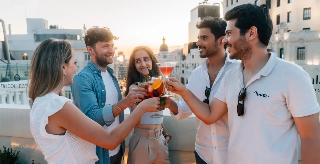 Five young friends toast with colorful cocktails on a rooftop bar in Madrid at sunset.