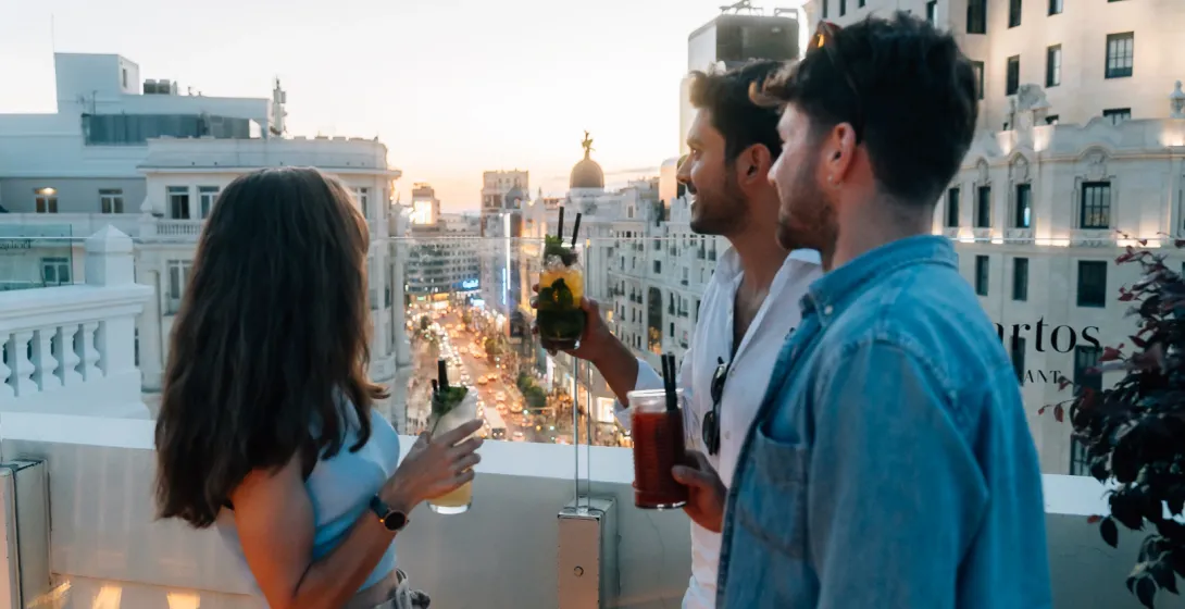 Three friends with cocktails on a rooftop terrace overlooking the busy streets of Madrid at sunset.