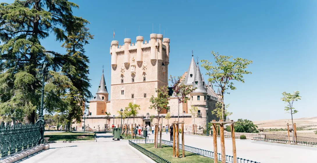 The Alcázar of Segovia, a stone castle with turrets, seen from a park on a sunny day.