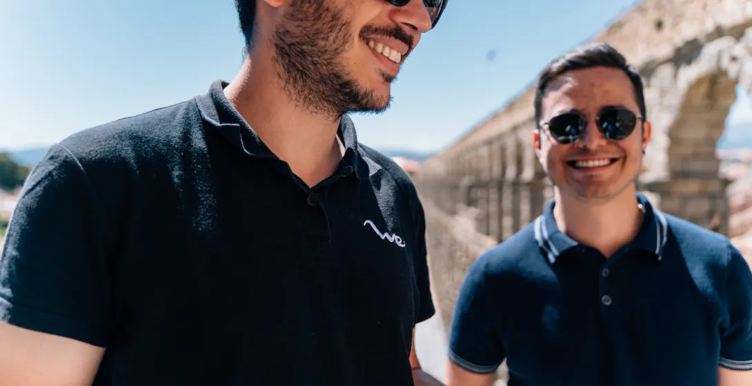 Two young men in sunglasses stand smiling in front of the Aqueduct of Segovia on a sunny day.