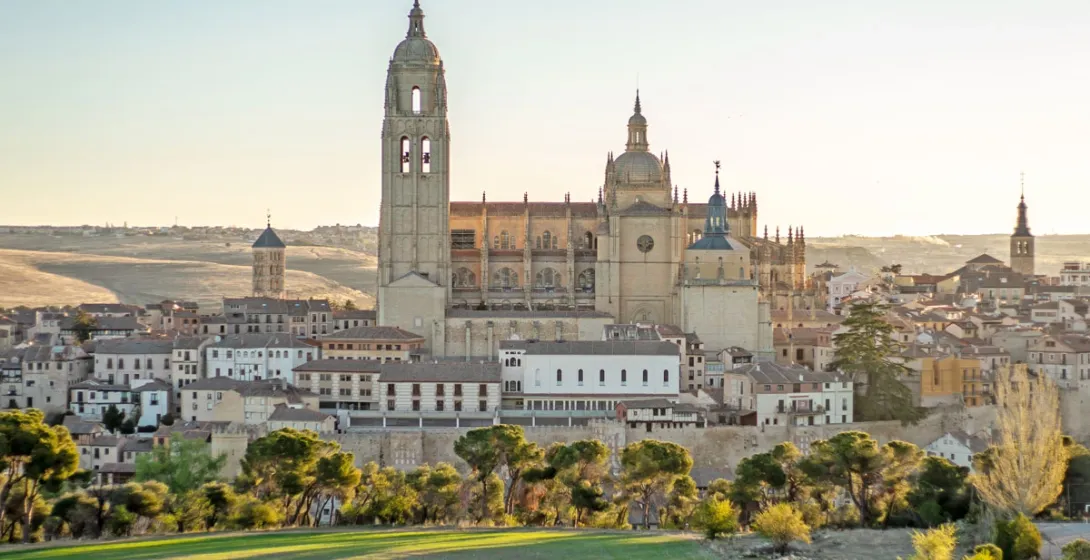 The Segovia Cathedral stands over the old town, seen from a green hill at sunset.