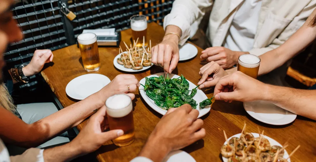 Friends share plates of tapas, including Padrón peppers, and glasses of beer at a wooden table in a bar.