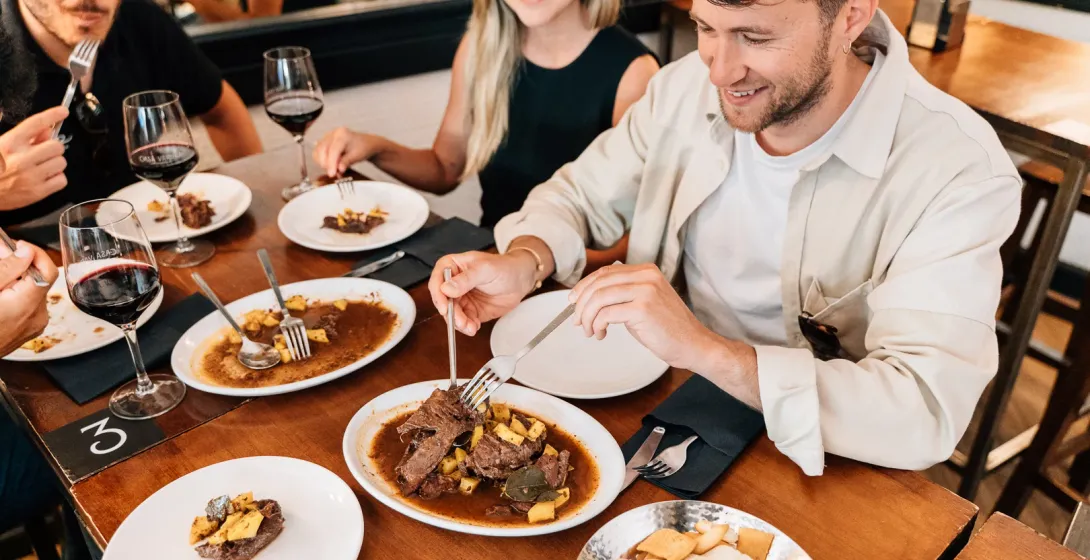 Three people at a wooden table sharing plates of beef stew and drinking glasses of red wine in a restaurant.