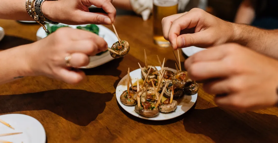 Hands picking up mushroom tapas with toothpicks from a plate on a wooden table.