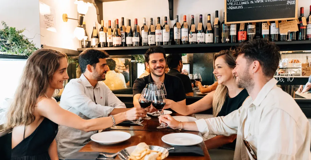 A group of five friends sits at a table in a bar, smiling and toasting with glasses of red wine.