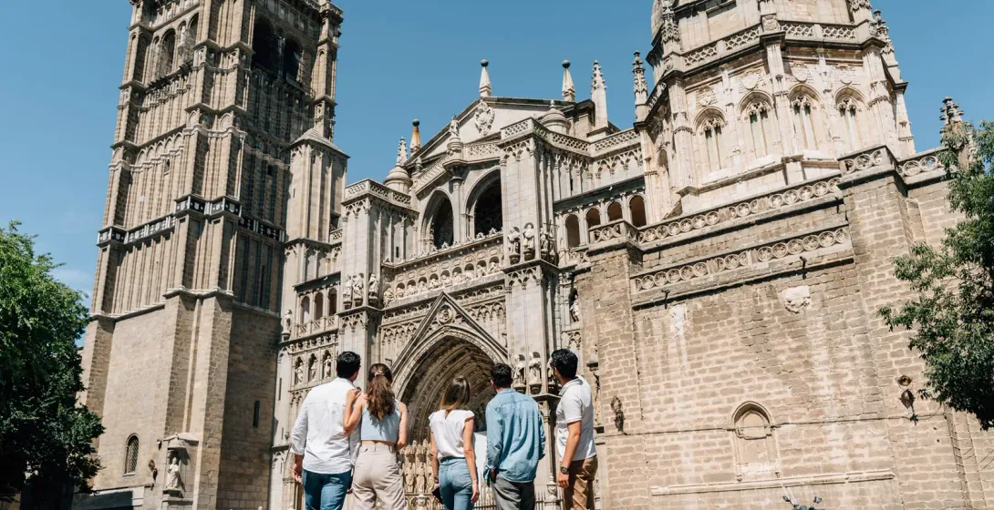 A group of five people, seen from behind, look up at the intricate stone facade of the Toledo Cathedral on a sunny day.