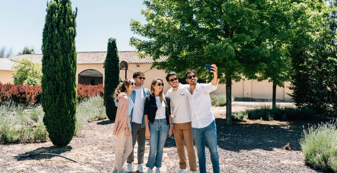 A group of five friends takes a smiling selfie in a sunny garden with cypress trees and a villa in the background.