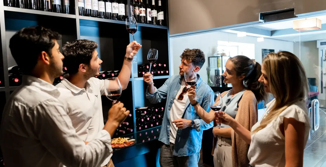 A group of people tasting red wine in a shop. One man holds his glass up to inspect the color against a wall of bottles.
