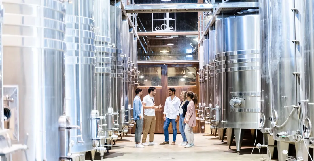 A group of people on a tour, standing between large stainless steel fermentation tanks in a modern winery.