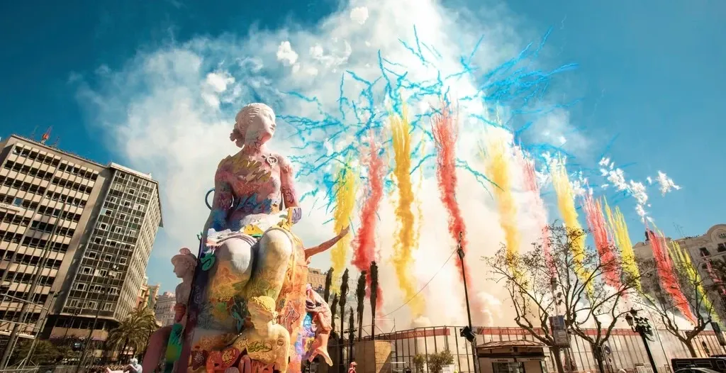A large, colourful 'falla' statue with a daytime firework display of coloured smoke exploding behind it in a city square.