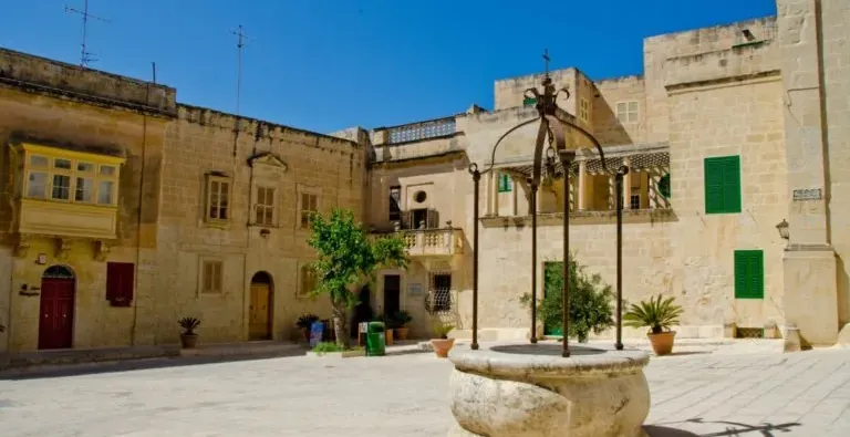 Empty stone square with a historic stone well in the foreground. Traditional buildings with balconies and colorful shutter...