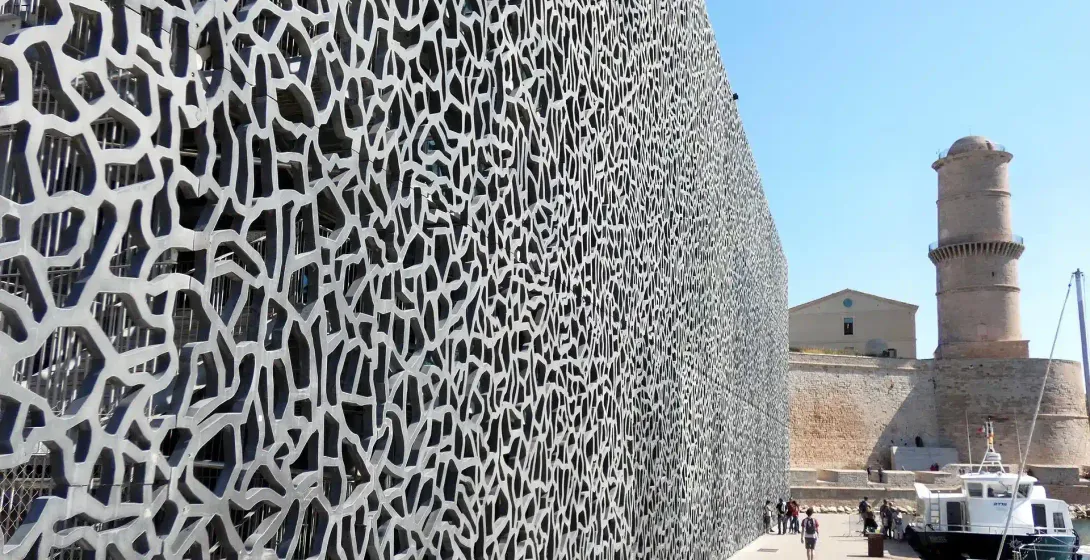 Modern building with intricate, net-like facade and old stone fort under a clear blue sky on a sunny day in Marseille.