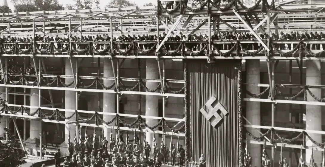 Black and white photo of a building under construction, decorated with garlands and a large swastika banner. People are ga...