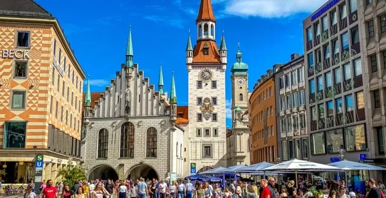Crowded Marienplatz in Munich with the Old Town Hall and the tower of the New Town Hall under a blue sky.