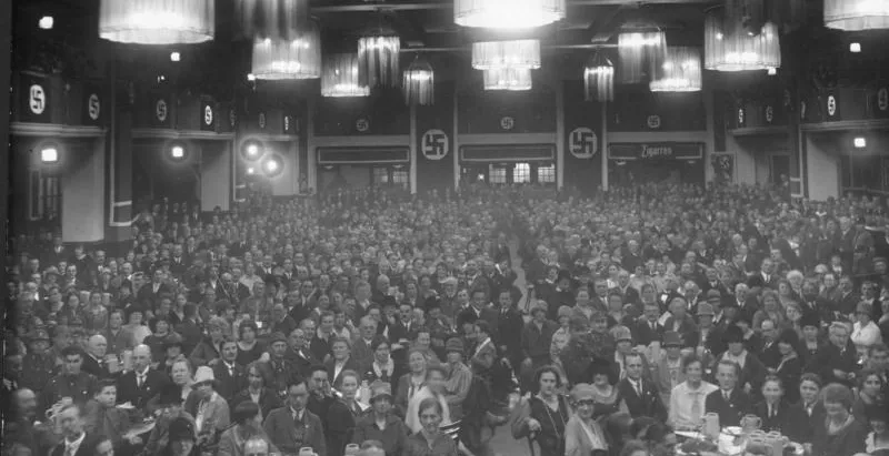Interior of a crowded Munich beer hall during the Nazi era with swastika banners and chandeliers.