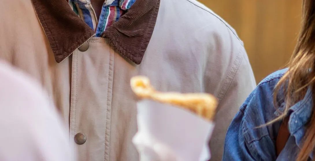 Smiling man watching a crepe being handed to a person in a denim jacket on a street.