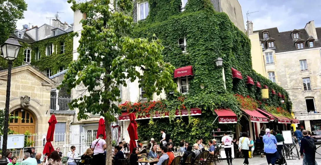 Building covered in green ivy and red awnings, with an outdoor restaurant patio in Paris. People are dining under a cloudy...