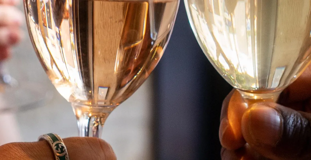 Two hands holding wine glasses, one with rosé wine and the other with white wine, clinking in a toast. One hand wears rings.