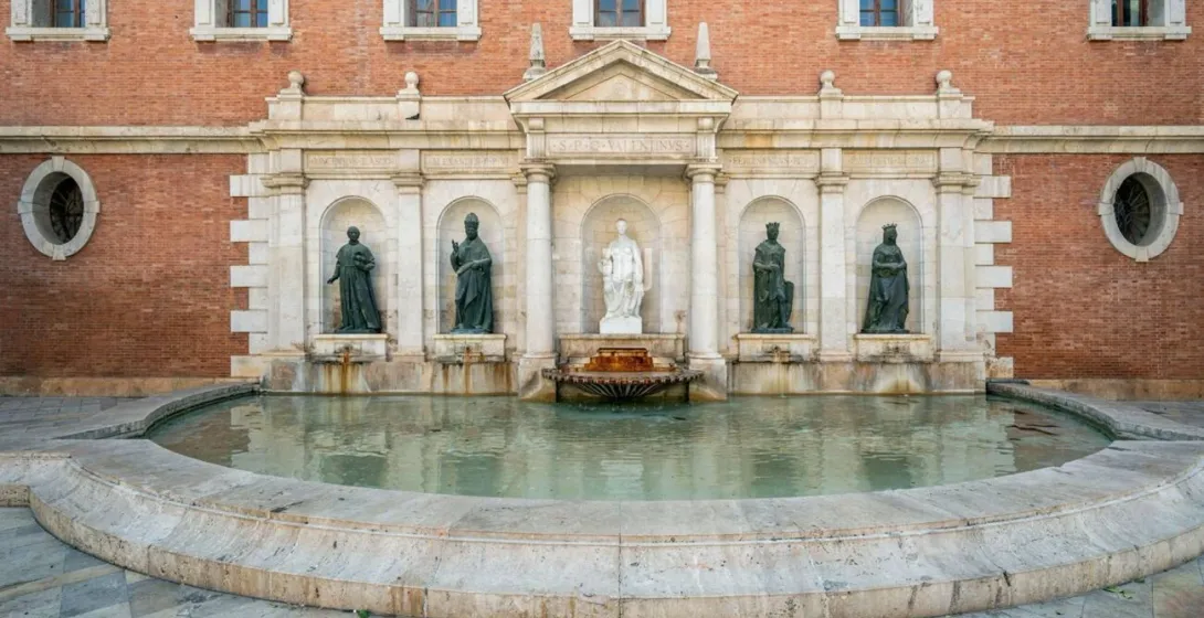 A wide, curved stone fountain in front of a brick building with statues in arched niches.
