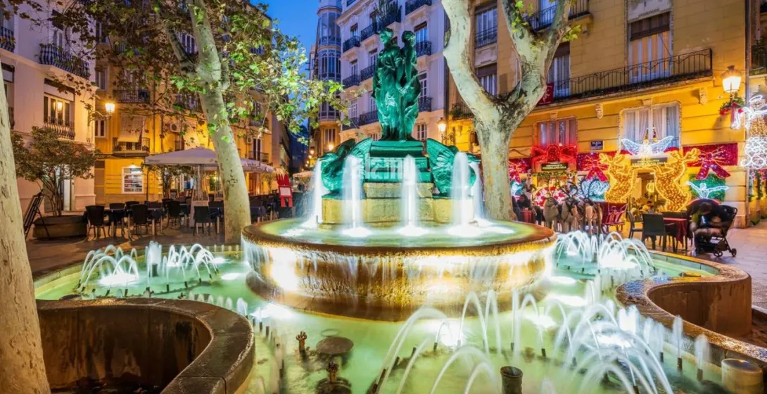 An illuminated fountain in Plaza Rodrigo Botet in Valencia, surrounded by trees and buildings with Christmas lights.