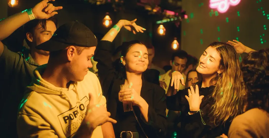 People dancing in a dimly lit club or bar, with green laser lights and a pink neon musical note sign.