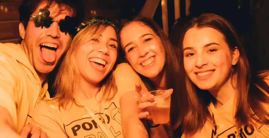 Four young people, two men and two women, smiling in a dimly lit bar. They wear yellow t-shirts with 'PORTO PUB CRAWL' and...
