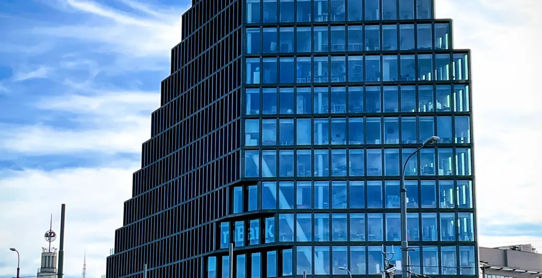 Modern Bałtyk building in Poznań with a stepped glass facade, set against a blue sky, with a street and historical tower v...
