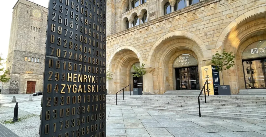 Dark monument with gold numbers and 'HENRYK ZYGALSKI' on a paved plaza, facing Poznan Imperial Castle with arches.