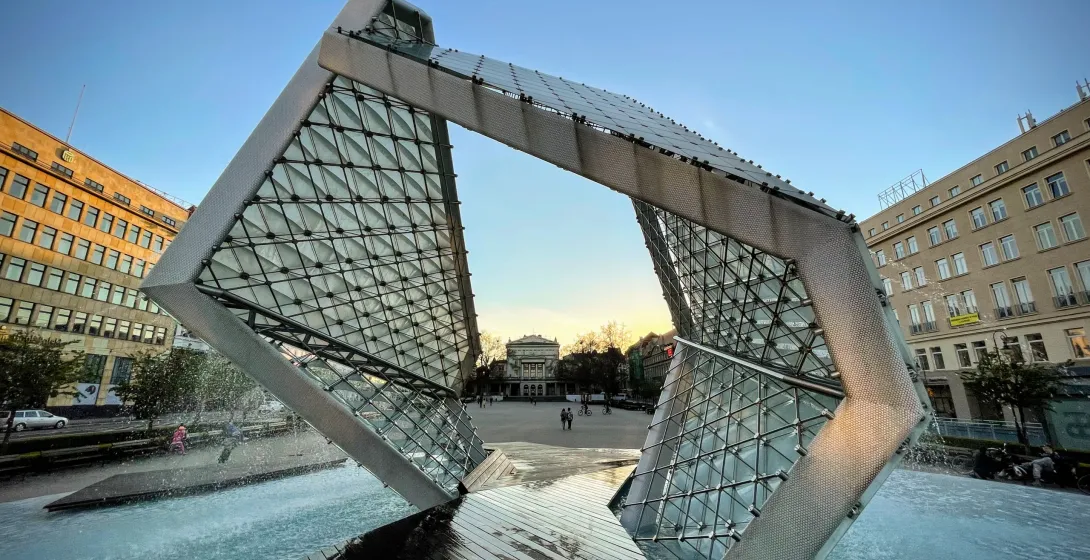 Modern glass and metal fountain with water jets, shaped like an open cube, in a city square at dusk. Buildings in the back...