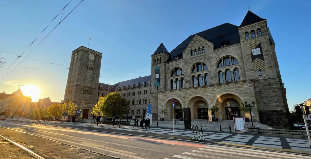 Stone Imperial Castle building in Poznań with a clock tower, tram tracks, and a crosswalk in the foreground. Sun is low on...