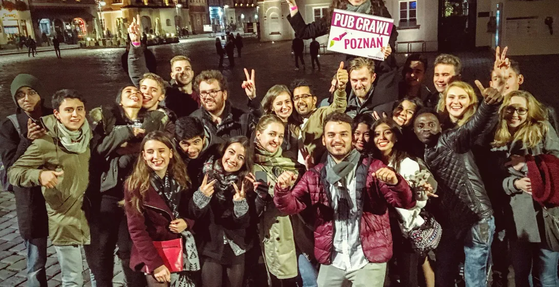 Large group of smiling young adults on a wet cobblestone street at night, one holding a 'PUB CRAWL POZNAN' sign, historic ...