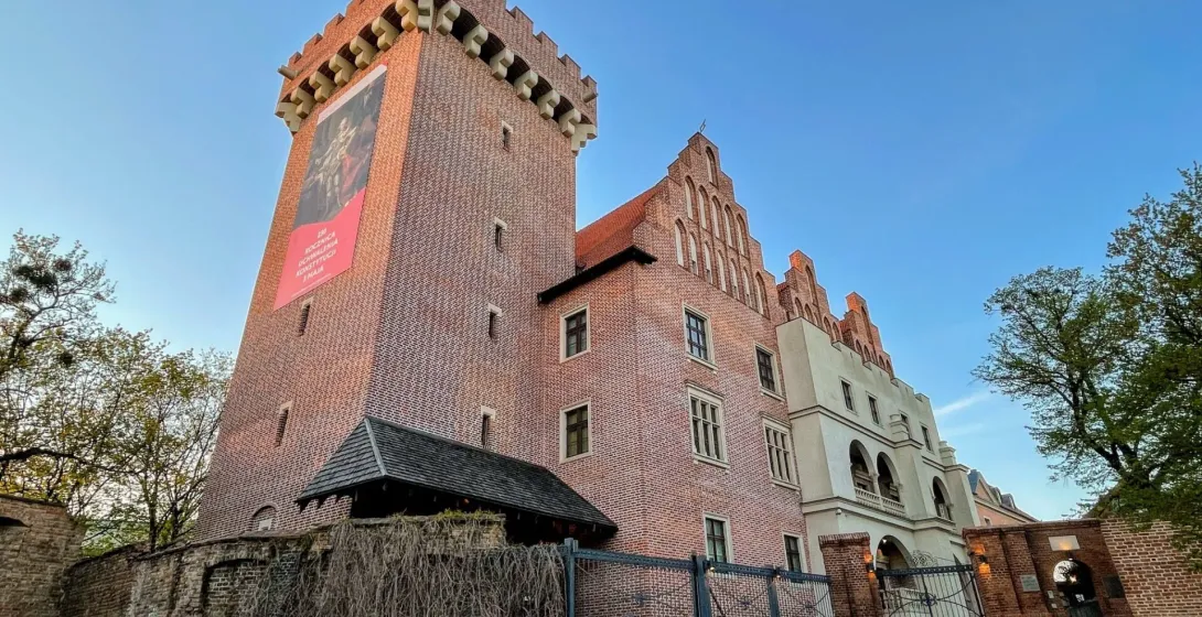 View of the Royal Castle in Poznań, a large brick building with a tall tower, gabled facade, and old wall, under a blue sky.