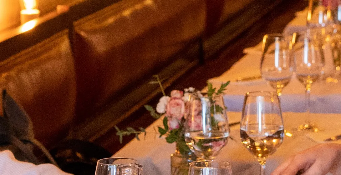 People dining at a restaurant table with bread, a water bottle, wine glasses, and a cheese board. Warm interior lighting.