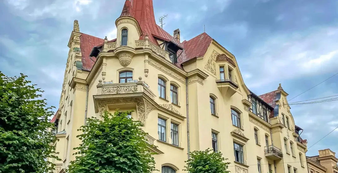 Ornate Art Nouveau building in Riga with a yellow facade, red roof, and central spire, framed by green trees against a clo...