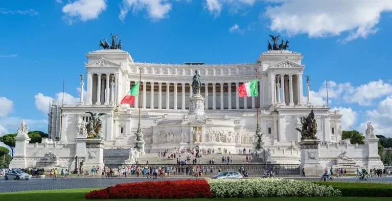 Grand white marble Altar of the Fatherland in Rome under a blue sky with clouds. Italian flags fly on either side. People ...