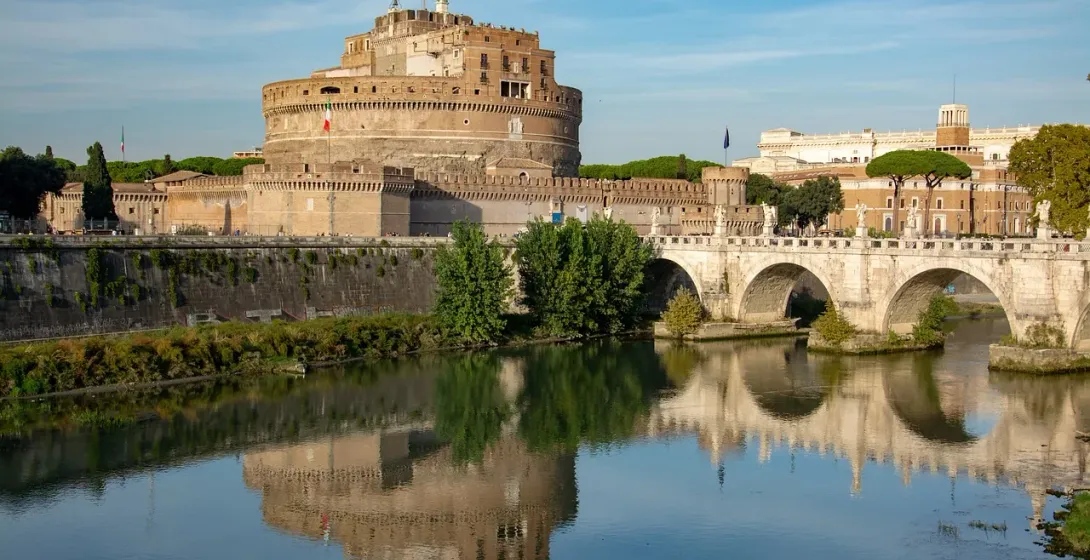 Castel Sant'Angelo and Ponte Sant'Angelo spanning the Tiber River under a blue sky, with reflections on the water.