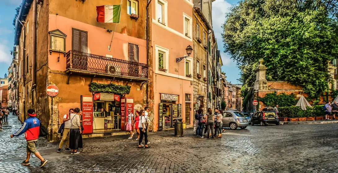 Cobblestone street in Rome with historic buildings, an Italian flag, people walking, and parked cars under a blue sky.
