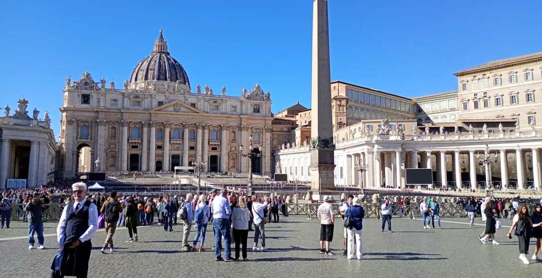 Tourists walking in St. Peter's Square with the central obelisk and St. Peter's Basilica facade behind.