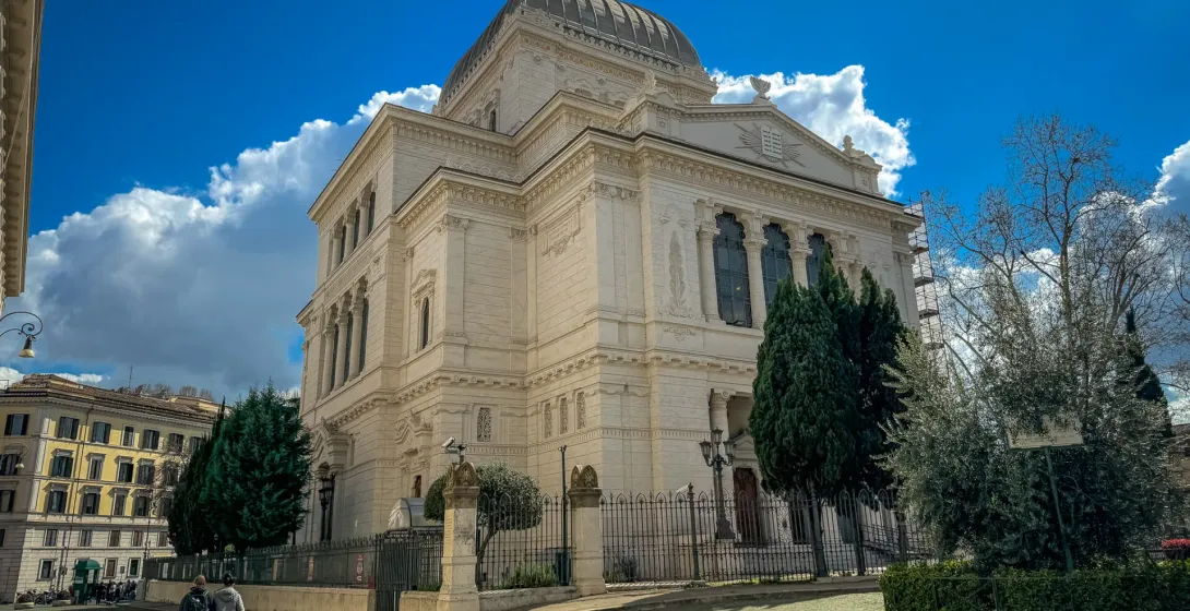 The Great Synagogue of Rome with its dome, a cobblestone street, green trees, and two people walking under a bright blue sky.