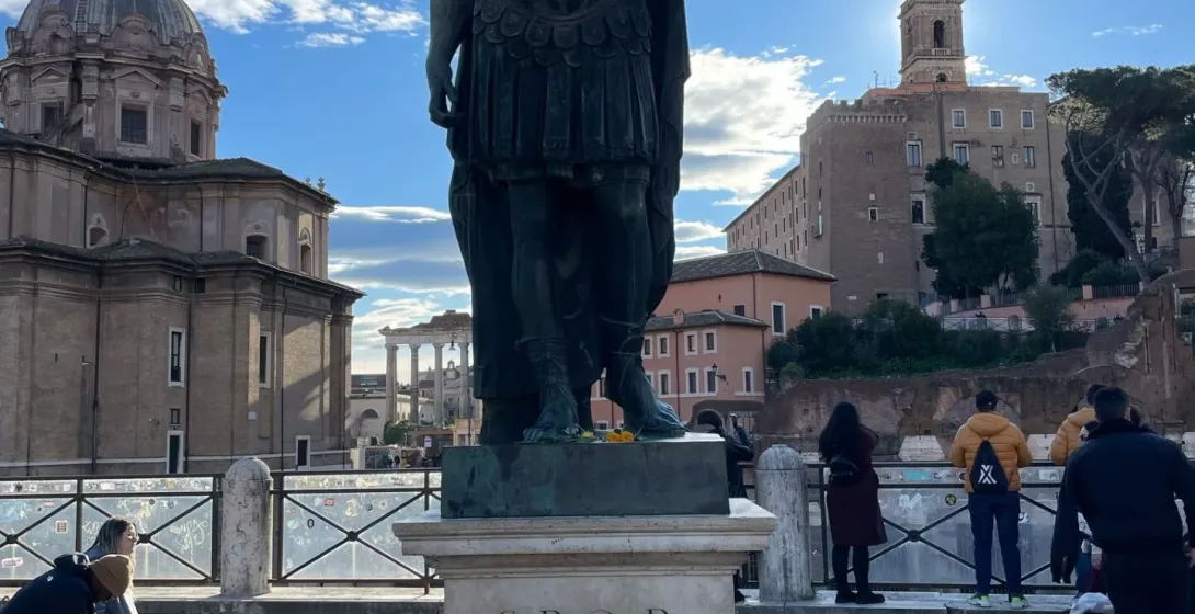 Bronze statue of Julius Caesar in Rome, with Roman Forum ruins and a domed church under a blue sky.