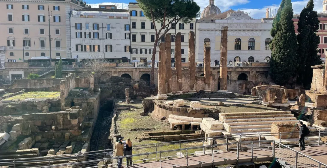 View of ancient Roman ruins with excavated levels, stone columns, and a modern wooden walkway. City buildings in background.