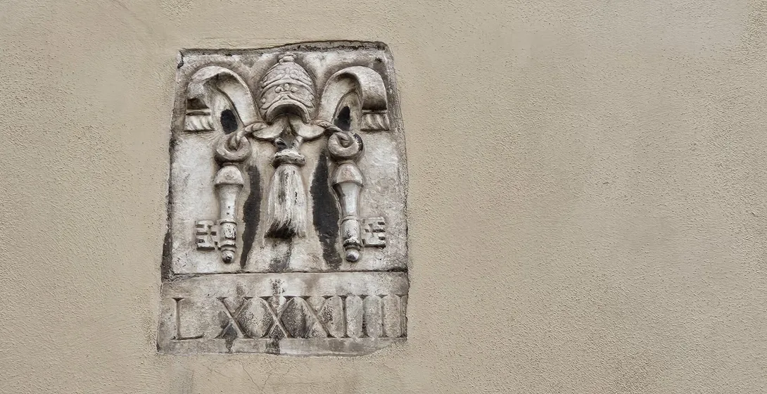 Stone relief on a textured wall showing a papal tiara, crossed keys, and tassels, with Roman numerals LXXXIII below.