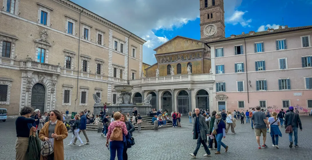Cobblestone Piazza Santa Maria in Trastevere, Rome, with a basilica, bell tower, fountain, and many people walking.