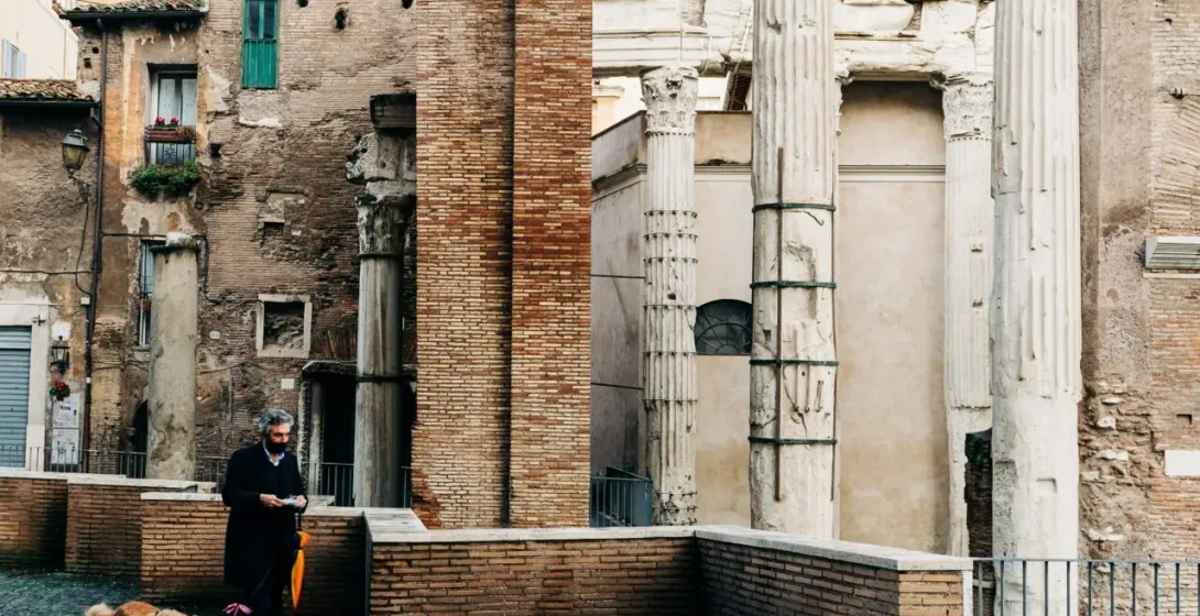 Man walking a golden retriever on a wet cobblestone street past ancient Roman ruins with tall columns and Latin inscriptions.