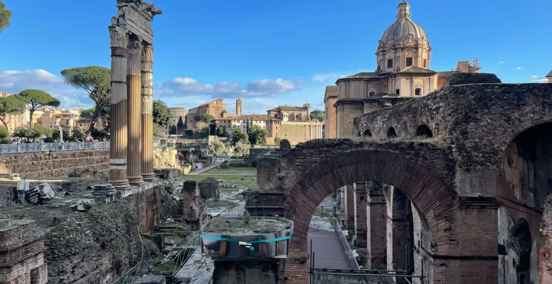 Ancient Roman Forum ruins with the Temple of Saturn columns, other temples, and scattered visitors under a blue sky.