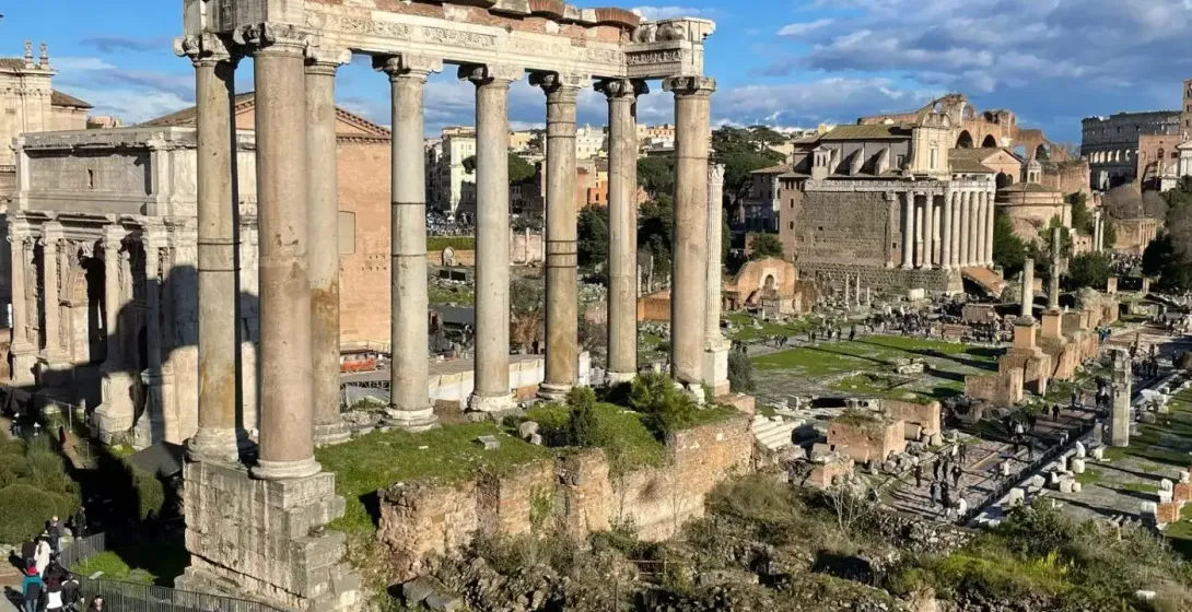 View of the Roman Forum in Rome, showing ancient ruins including eight tall columns of the Temple of Saturn under a blue sky.