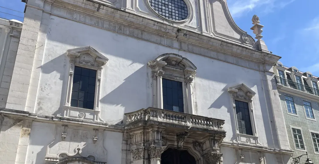 Ornate white facade of São Domingos Church in Lisbon, with large dark doors, a balcony, and a cross on the roof. People ga...