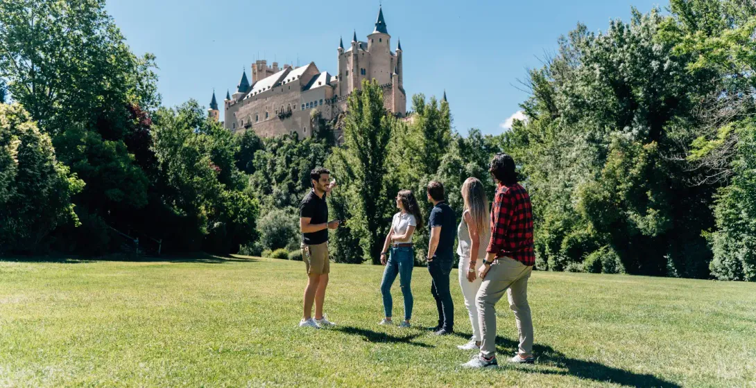 A tour guide speaks to a small group in a park with the Alcázar of Segovia castle in the background.
