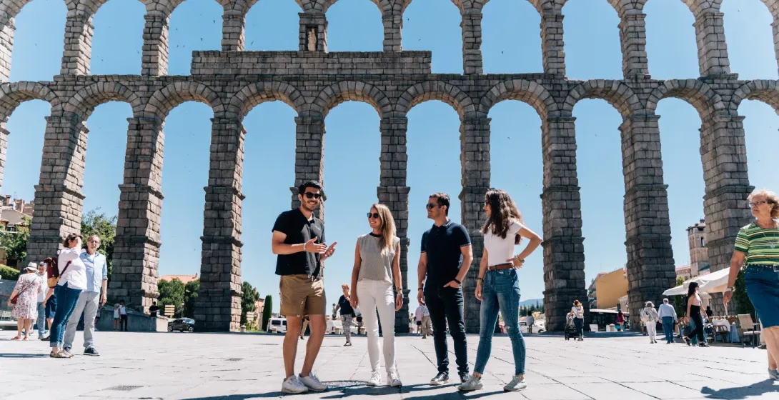 A tour guide talks to three tourists in front of the large, sunny Aqueduct of Segovia in Spain.