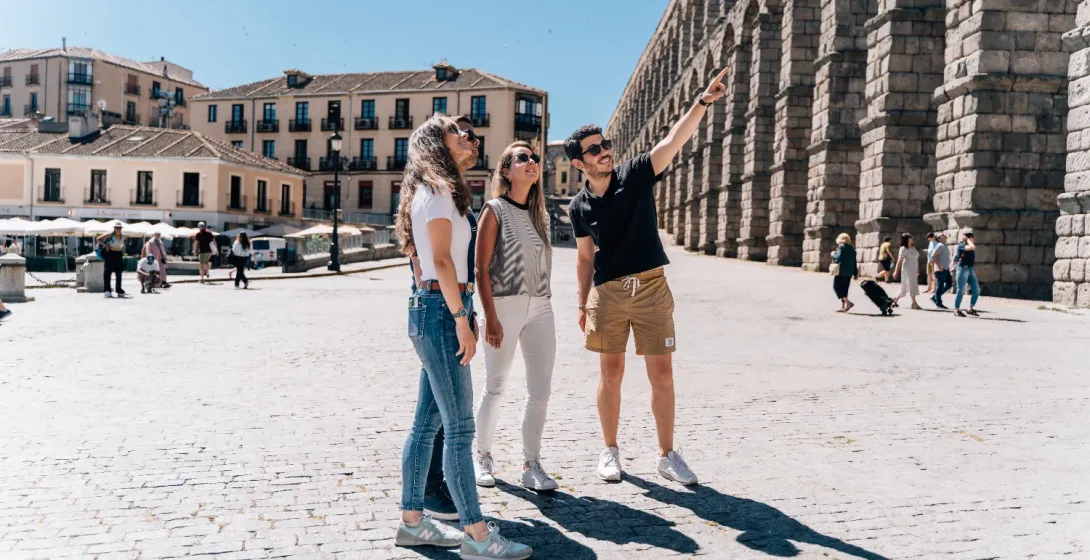 A tour guide points up at the Aqueduct of Segovia for three tourists on a sunny day.